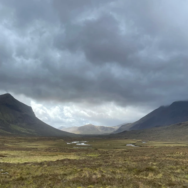 A view southwards along Glen Sligachan, a wide flat-bottomed valley with mountains along each side. A small river winds along the valley floor through gorse and scrub. There are dark clouds in the sky, but at the far end of the valley a mountain is in sunlight.