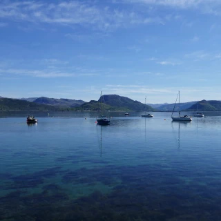 A view across Loch Carron from Plockton. The water is clear, with seaweed visible on the loch bed. Four small sailing boats with their sails furled and two smaller boats are at anchor. In the distance are wooded hills, under a blue sky with a few wisps of cloud.