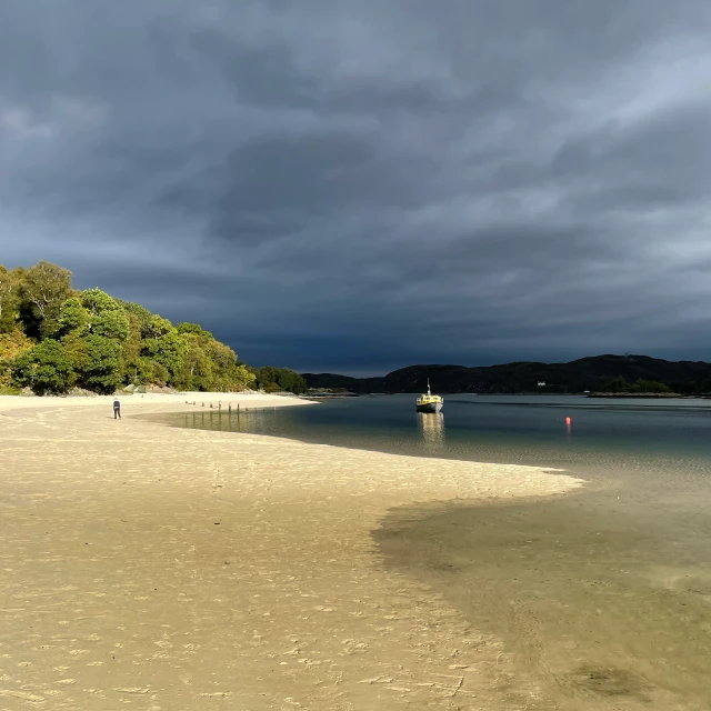 A view along Morar Bay, towards the sea. There are dark clouds in the sky, and the hills on the far side of the bay are almost black, but the sand and trees in the foreground are brightly lit. The sunlight highlights a small boat floating on the dark water.