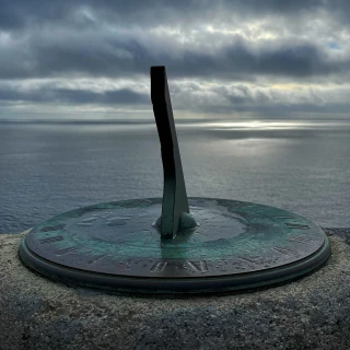 A metal sundial with a blue-green patina, mounted on a granite disc. Beyond the sundial is the sea, and while the sky is almost overcast a small patch of the sea is sunlit.