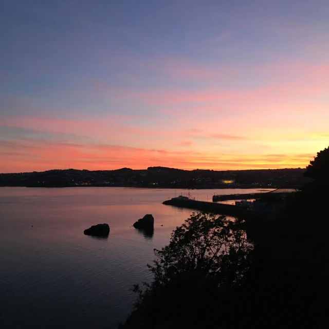 A view across Torbay. The sun has just set, and the sky is coloured red, yellow, blue, and purple. The far side of the bay, the harbour walls, two rocky islands, and some nearby trees are silhouetted against the sky and its reflection in the calm water.